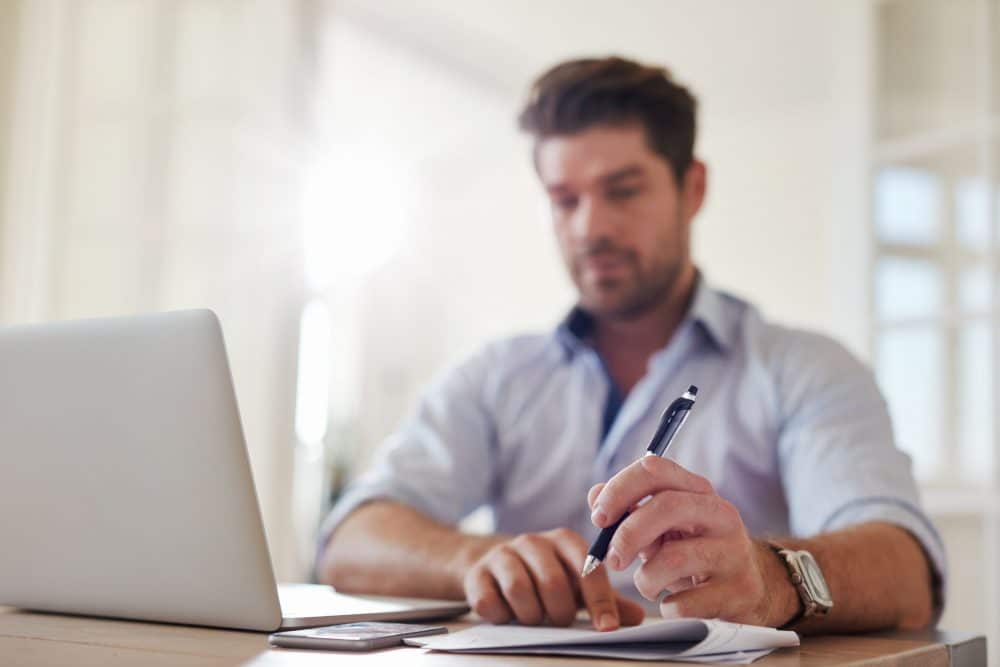 Person writing on a pad of paper with their laptop open in front of them.