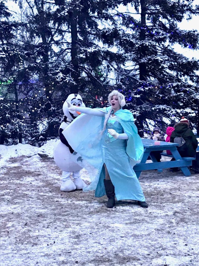 Elsa with blond hair wearing a blue dress, cloak and earmuffs sings. An Olaf snowman costumed character stands behind her.