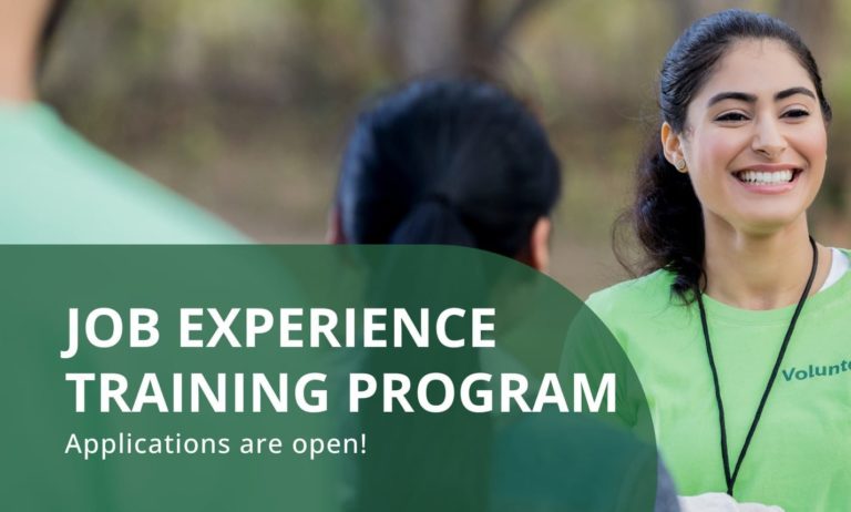 A young women smiles facing towards gathered people just out of focus. The text "Job Experience Training Program, applications are open" appears in the bottom left.